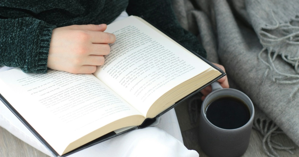 Person hält ein aufgeschlagenes Buch und eine Tasse Kaffee in den Händen